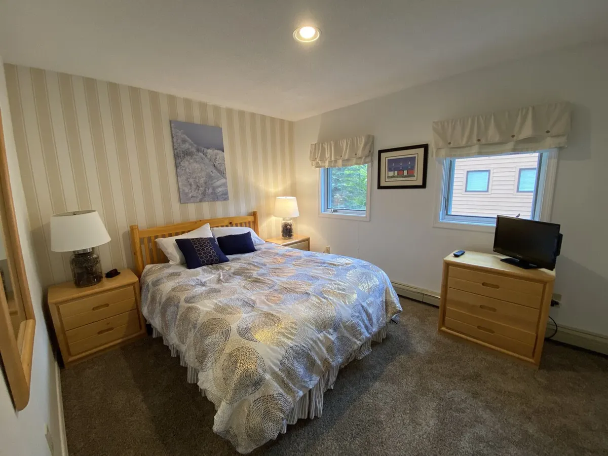A calm, restful bedroom featuring a plush queen bed with neatly arranged bedding, nightstands on either side, a window offering gentle natural light, and a TV mounted for relaxation. The room exudes a peaceful atmosphere perfect for restful nights.