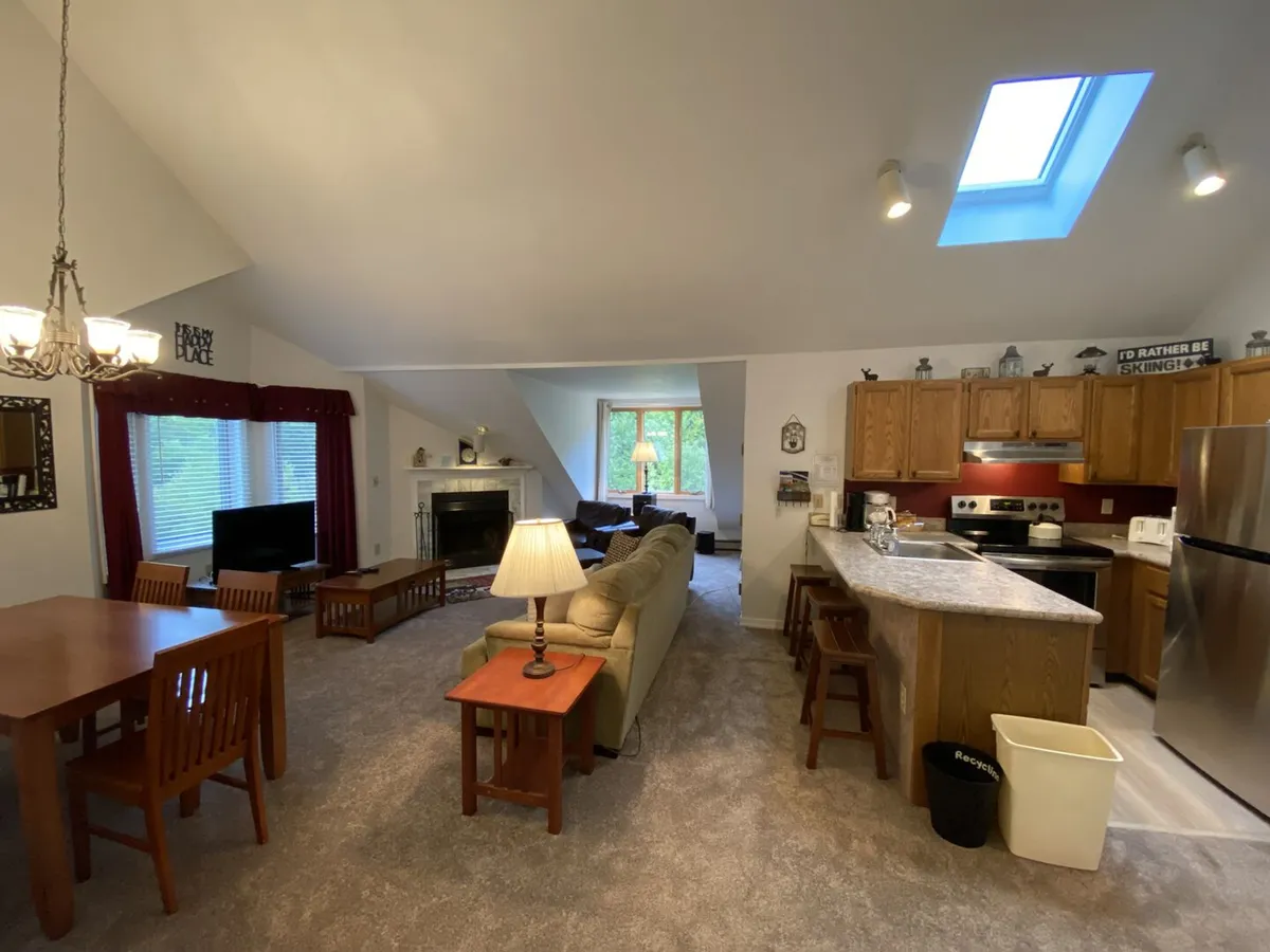 This inviting condo interior features a sunlit open-concept living area with a modern sofa, warming fireplace, bright skylight, dining table perfect for family meals or game nights, and a contemporary kitchen island ideal for gathering and breakfast.