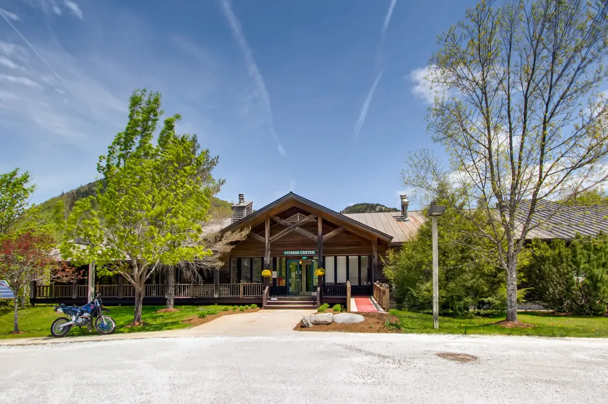 The image showcases the property's main entrance framed by a landscaped garden, with decorative planters adding a charming touch. The mountain backdrop connects the property to the surrounding natural environment, suggesting outdoor adventures and community gatherings just beyond the doorstep.