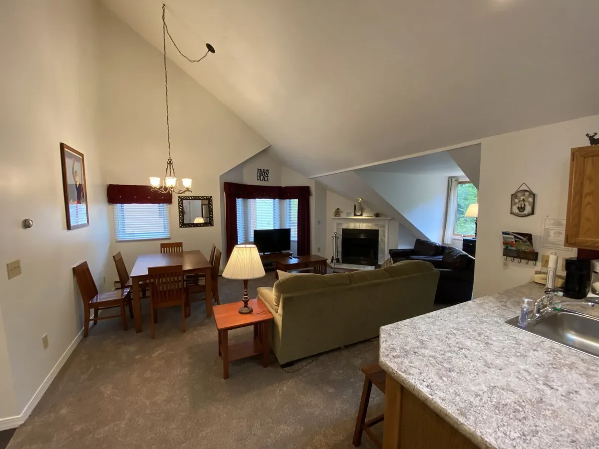 This image showcases an open-plan living area featuring a dining table, comfortable sofa, a fireplace, and the kitchen counter in the background. The warm color palette and visible seating arrangements suggest a space perfect for both relaxation and entertaining guests.