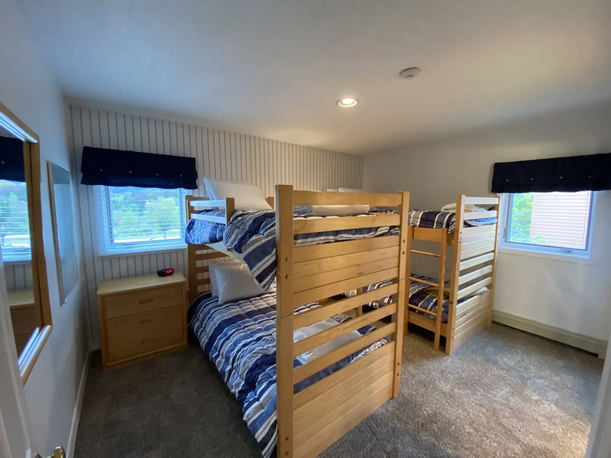 A lively and friendly bedroom with two sets of bunk beds, cheerful bedding, windows providing natural sunlight, a dresser for storage, and a mirror. The setup is ideal for kids or friends to share stories and enjoy cozy sleepovers.
